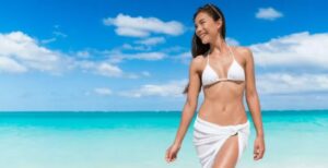 A woman in white bikini and towel walking on the beach.