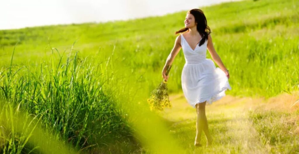 A woman in white dress holding flowers walking through grass.